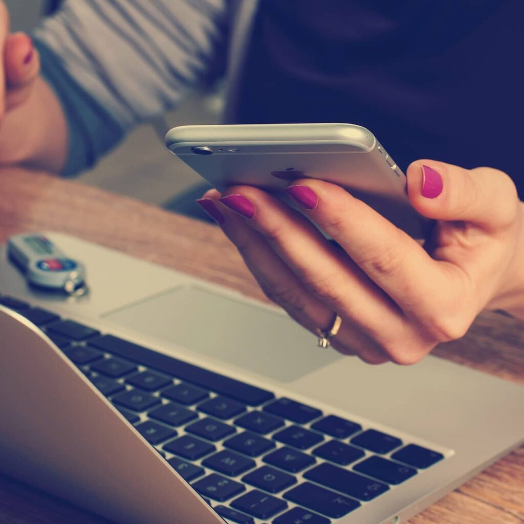 Kobieca dłoń z telefonem w ręku, siedząca przy laptopie (czy warto założyć własny biznes)